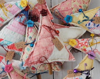 May include: A collection of fabric Christmas tree ornaments made from quilted patchwork scraps in various pastel and floral patterns. Each triangular ornament features a cinnamon stick trunk, a small button at the top, and a brown paper tag labeled Joy or Noel.