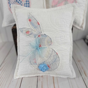 May include: White quilted pillow with a bunny applique. The bunny is made of patterned fabric with blue and pink floral designs. A blue ribbon and a small blue fabric flower are attached to the pillow.