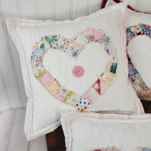 May include: A square white quilted pillow featuring a central heart shape constructed from various small, colorful patterned fabric scraps. A single pink button is sewn into the center of the heart. The pillow has a light pink fabric border.