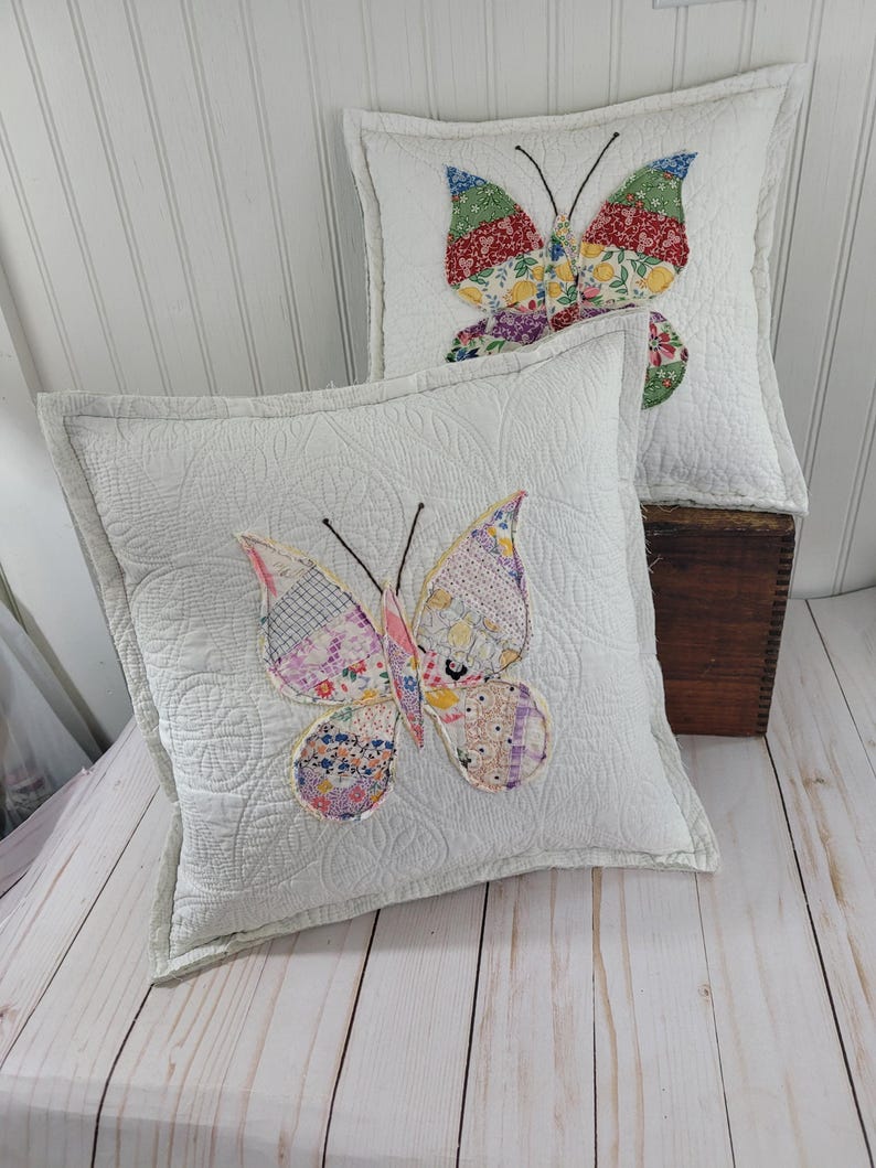 Op de afbeelding: Twee witte vierkante sierkussens met gewatteerde stiksels en vlinderappliqu&eacute;s. De vlindervleugels zijn gemaakt van kleurrijke patchworkstof. De kussens liggen op een licht houten oppervlak.