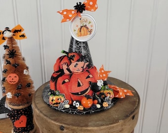 May include: A Halloween-themed witch hat with a pumpkin and black cat design. The hat features an orange polka dot bow and a circular emblem with a pumpkin and the word "Boo!". The hat is decorated with small pumpkins and spiders.