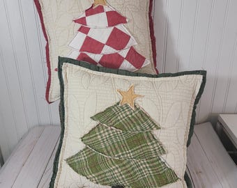 May include: Two square quilted throw pillows with cream-colored fabric featuring a stitched leaf pattern. One pillow displays a red and white checkered Christmas tree, while the other features a green plaid Christmas tree, both topped with a yellow star.