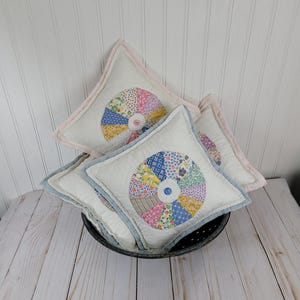 May include: Three handmade quilted pillow covers with a colorful patchwork design in the shape of a pinwheel. The pillows are in a metal bowl on a white wood surface.