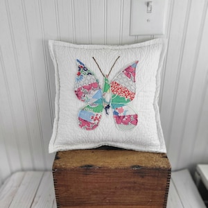 Op de afbeelding: Een vierkant wit kussen met een gewatteerd patroon, versierd met een kleurrijke vlinderapplicatie. De vlindervleugels zijn gemaakt van diverse stoffen met patronen in roze, blauw, groen en rood. Het kussen ligt op een houten kist.