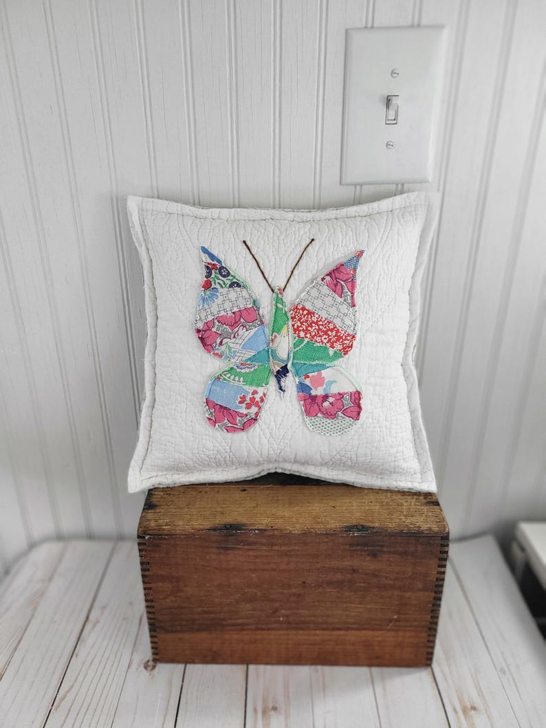 Op de afbeelding: Een vierkant wit kussen met een gewatteerd patroon, versierd met een kleurrijke vlinderapplicatie. De vlindervleugels zijn gemaakt van diverse stoffen met patronen in roze, blauw, groen en rood. Het kussen ligt op een houten kist.