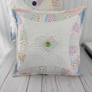 May include: Two square, quilted pillows with a patchwork design. Each pillow features a white center with a heart-shaped appliqué and a button. The edges are trimmed with pastel fabrics. The pillows rest on a light-colored wooden surface.