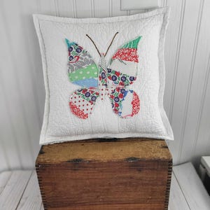 Op de afbeelding: Een vierkant wit gewatteerd kussen met een kleurrijke vlinderapplicatie. De vlinder heeft vleugels met rood, blauw, groen en grijs patroonstof. Het kussen ligt op een houten kist.