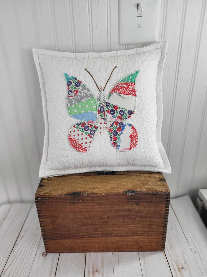 Op de afbeelding: Een vierkant wit gewatteerd kussen met een kleurrijke vlinderapplicatie. De vlinder heeft vleugels met rood, blauw, groen en grijs patroonstof. Het kussen ligt op een houten kist.