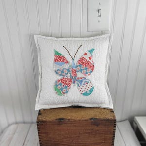 Op de afbeelding: Een vierkant, wit gewatteerd kussen met een kleurrijke vlinderapplicatie. De vlindervleugels hebben rood, blauw en groen bedrukte stoffen. Het kussen ligt op een houten kist, met een witte muur en een lichtschakelaar op de achtergrond.