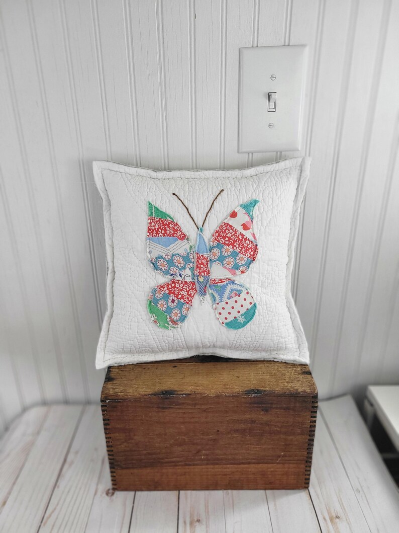 Op de afbeelding: Een vierkant, wit gewatteerd kussen met een kleurrijke vlinderapplicatie. De vlindervleugels hebben rood, blauw en groen bedrukte stoffen. Het kussen ligt op een houten kist, met een witte muur en een lichtschakelaar op de achtergrond.