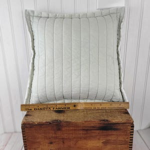 Op de afbeelding: Een vierkant, lichtblauw gewatteerd kussen met verticale stiksels. Het kussen ligt op een houten kist. Een houten liniaal met de tekst "The DAKOTA FARMER" ligt op de kist. Het kussen is ongeveer 35,5 cm lang.