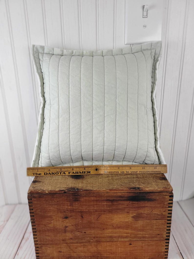 Op de afbeelding: Een vierkant, lichtblauw gewatteerd kussen met verticale stiksels. Het kussen ligt op een houten kist. Een houten liniaal met de tekst "The DAKOTA FARMER" ligt op de kist. Het kussen is ongeveer 35,5 cm lang.