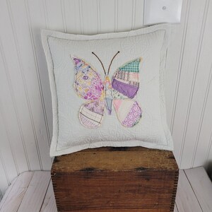 Op de afbeelding: Een vierkant, wit gewatteerd kussen met een patchwork vlinderontwerp in tinten paars, roze en groen. De vlinder heeft bruine antennes en is gecentreerd op het kussen. Het kussen is geplaatst op een houten kist.
