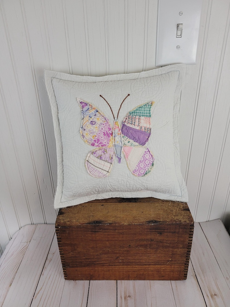 Op de afbeelding: Een vierkant, wit gewatteerd kussen met een patchwork vlinderontwerp in tinten paars, roze en groen. De vlinder heeft bruine antennes en is gecentreerd op het kussen. Het kussen is geplaatst op een houten kist.