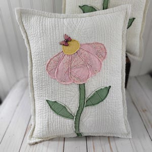 May include: A rectangular white quilted pillow featuring a central fabric applique of a pink flower with a yellow center and a small purple butterfly on top. The flower has a green stem and two green leaves, all stitched onto the white fabric.