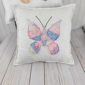 May include: A square white quilted throw pillow featuring a central butterfly appliqué made from various floral and patterned fabric patches in shades of pink, blue, and yellow. The butterfly has two thin black embroidered antennae.