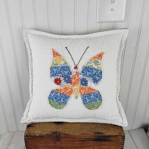 Op de afbeelding: Een vierkant, wit gewatteerd kussen met een kleurrijke vlinderapplicatie. De vlinder heeft oranje, blauwe en groene vleugels met patronen en een zwart lichaam. Het kussen staat op een houten kist, wat een rustieke uitstraling cre&euml;ert.