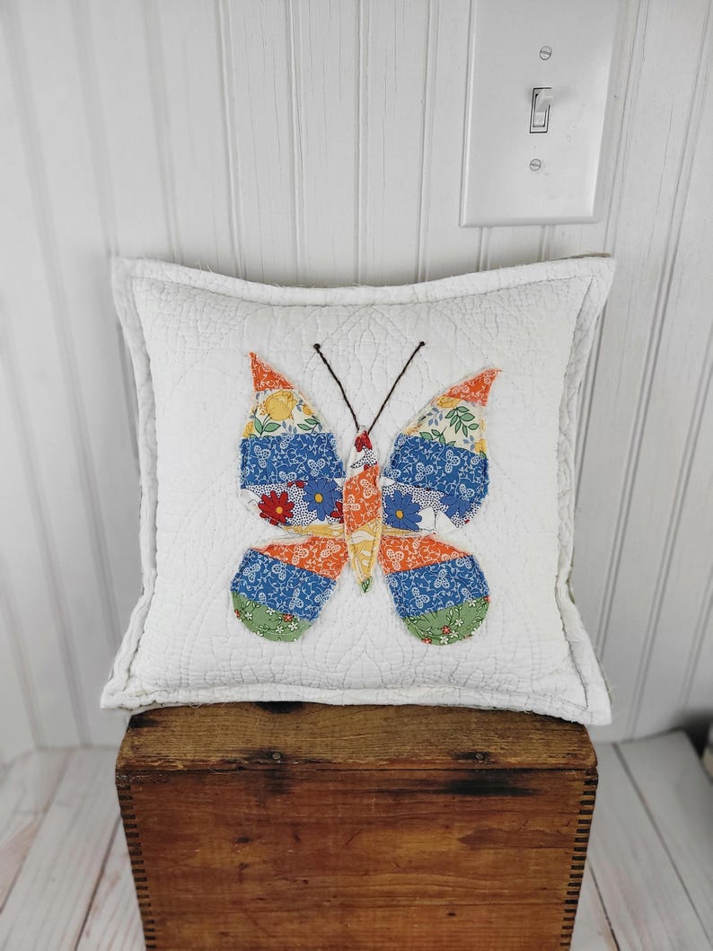 Op de afbeelding: Een vierkant, wit gewatteerd kussen met een kleurrijke vlinderapplicatie. De vlinder heeft oranje, blauwe en groene vleugels met patronen en een zwart lichaam. Het kussen staat op een houten kist, wat een rustieke uitstraling cre&euml;ert.