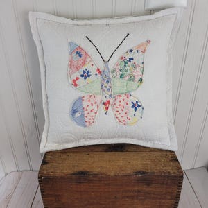 Op de afbeelding: Een vierkant wit kussen met een gewatteerd patroon en een kleurrijke vlinderapplicatie. De vlindervleugels zijn gemaakt van verschillende stoffen met bloemmotieven in tinten rood, blauw en groen. Het kussen ligt op een houten kist.