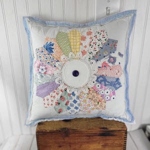 May include: A square white throw pillow featuring a central sunburst pattern made of various colorful fabric scraps in floral, plaid, and geometric prints. A white lace doily sits in the center with a single dark purple button and light blue trim.