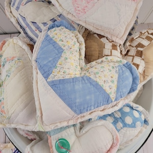 May include: A collection of handmade heart-shaped quilted items in various fabrics and patterns. The hearts feature patchwork designs, floral prints, stripes, and polka dots, with a mix of pastel and neutral colors. Some hearts have a button detail.