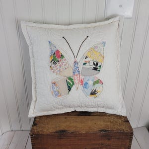 Op de afbeelding: Een vierkant, wit gewatteerd kussen met een kleurrijk patchwork vlinderontwerp. De vlindervleugels hebben verschillende bloemen- en patronenstoffen in tinten blauw, roze, geel en groen. Het kussen ligt op een houten kist.