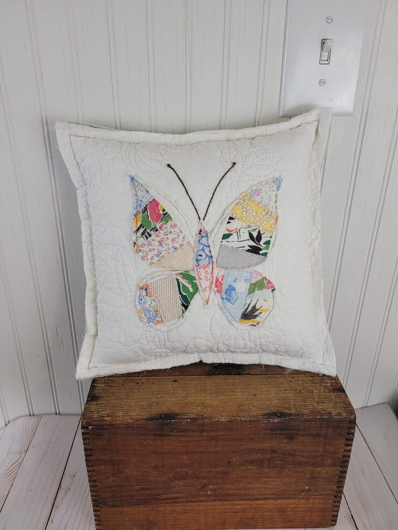 Op de afbeelding: Een vierkant, wit gewatteerd kussen met een kleurrijk patchwork vlinderontwerp. De vlindervleugels hebben verschillende bloemen- en patronenstoffen in tinten blauw, roze, geel en groen. Het kussen ligt op een houten kist.