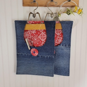 May include: Two denim fabric storage bags with red bandana-patterned pockets and decorative red fabric buttons. The bags hang from vintage metal hangers attached to a wooden board. A small wooden clothespin is attached to one bag.