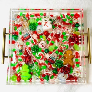 May include: A clear acrylic tray filled with a festive assortment of Christmas candies. The tray is adorned with red, green, and white candies, including candy canes, gingerbread men, and wrapped chocolates. Gold handles are attached to the sides.