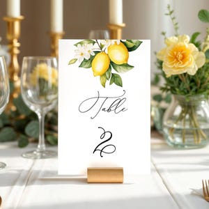 Puede incluir: Una tarjeta blanca con una ilustración de limón y el texto "Mesa 2" impreso en ella. La tarjeta está en un soporte dorado y está sobre una mesa blanca con un mantel blanco.