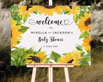 May include: A rectangular white sign featuring a watercolor sunflower border and black text that reads: welcome to MIRELLA AND JACKSON'S Baby Shower 5 JUNE 2021. The sign is displayed on a light-colored wooden easel stand.