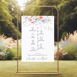 May include: A wedding timeline sign with a floral design in shades of pink, purple, and orange. The sign is displayed on a gold metal stand and includes the wedding schedule, date, and names of the couple.