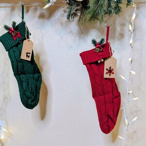 May include: Two cable-knit Christmas stockings, one green and one red, hang from metal hooks. Each stocking has a decorative tag and is adorned with faux berries and pine. Festive holiday decor.