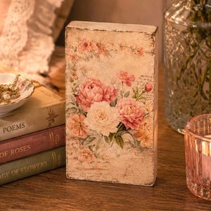 Puede incluir: Un bloque rectangular de madera con un acabado envejecido de color crema y un diseño floral impreso de peonías rosas y blancas. El bloque muestra texto de estilo vintage e ilustraciones botánicas en su superficie frontal.