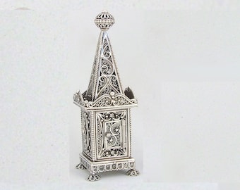Op de afbeelding: Een zilveren filigraandoos met een torenachtige top en sierlijke details. De doos heeft een rechthoekige basis met decoratieve voetjes en ingewikkelde patronen. De bovenkant heeft een puntig ontwerp met een kleine, ronde knop.