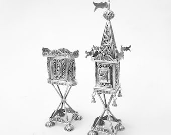 May include: Two ornate silver filigree structures. One is a small box on a stand, the other is a tall, tower-like structure with a flag on top and bells. Both have intricate openwork designs and are on a white background.