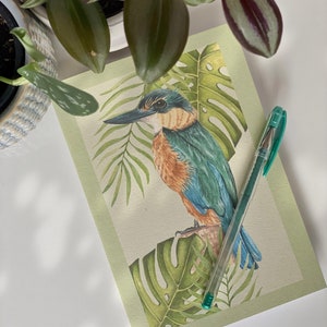 Peut inclure: Un oiseau martin-pêcheur vert et bleu perché sur une branche avec des feuilles tropicales vertes. Un stylo vert est posé sur la page.