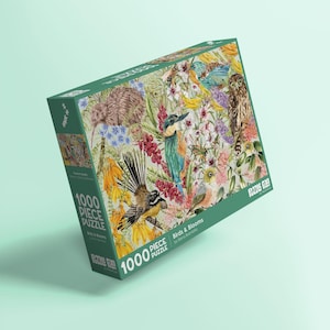 Może przedstawiać: Zielone pudełko z puzzlami z kolorową ilustracją ptaków i kwiatów. Na pudełku widnieje napis "1000 Piece Puzzle" i "Birds & Blooms". Grafika zawiera różne gatunki ptaków i żywe elementy kwiatowe.