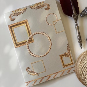 Puede incluir: Una cubierta de diario gris claro con un diseño decorativo de cuerda dorada, marcos y hojas. La cubierta tiene un acabado texturizado sutil.