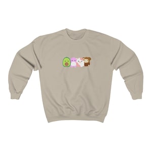 Puede incluir: Una sudadera beige claro con un gráfico de un aguacate, una criatura rosa, un gato blanco y un mono marrón.