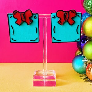 Puede incluir: Un par de pendientes de color azul turquesa con forma de cajas de regalo con lazos rojos. Los pendientes están en un soporte de acrílico transparente.