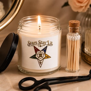 Puede incluir: Una vela encendida en un frasco de vidrio transparente con tapa negra, con una etiqueta blanca que dice Stars Stay Lit y un emblema de estrella de cinco puntas de colores. Al lado hay un frasco pequeño con cerillas y un cortamechas de metal negro.