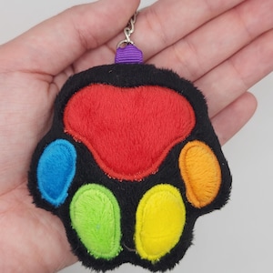 May include: A plush paw-shaped keychain with a black base and colorful pads. The central pad is red, with blue, green, yellow, and orange pads. A purple ribbon and silver keychain ring are attached.