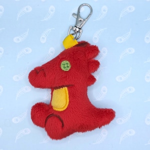 May include: A red plush dragon keychain with a yellow belly and green eye detail. The dragon has a silver metal clasp and a yellow fabric loop for attachment. The background is light blue with a feather pattern.