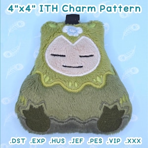 May include: A plush charm in the shape of a cartoon character, approximately 4 inches by 4 inches. The charm is primarily green with a cream-colored face and embroidered details. The text "4"x4" ITH Charm Pattern" is at the top, and file type extensions are at the bottom.