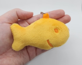 May include: A plush, yellow fish-shaped keychain. The fish has an orange eye and a smiling mouth. The keychain has a small orange fin on top and a silver metal ring for attaching to keys or a bag.