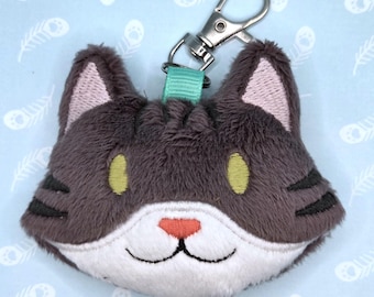 May include: A plush cat head keychain featuring dark gray fur, a white muzzle, and a coral-colored nose. The cat has embroidered pale green eyes, black whisker markings, and pink inner ears. A teal ribbon loop attaches the plush to a silver metal clasp.