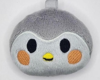 May include: A round, plush gray bird character with a soft texture, featuring two black embroidered eyes, a small yellow beak, and two orange circular patches on the cheeks. A small fabric loop is attached to the top of the plush item.