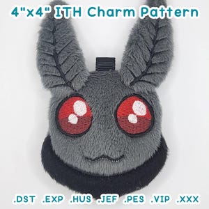 May include: A grey plush charm, approximately 10 cm x 10 cm, featuring a creature with large ears and red eyes, and a black trim. The text "4"x4" ITH Charm Pattern" is at the top. The bottom of the image shows file extensions .DST .EXP .HUS .JEF .PES .VIP .XXX.