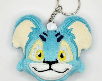 May include: A plush keychain featuring a cartoon cat face with blue fur, cream-colored ears, and yellow eyes. The cat has a smiling mouth and black whiskers. The keychain includes a silver metal ring and chain.